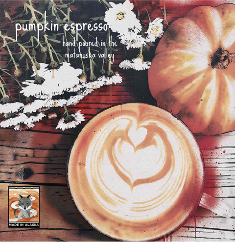 A square product label featuring a cup of latte art coffee on a rustic wooden table, with a whole pumpkin, white flowers, and overlaid text reading 'pumpkin espresso' and 'hand poured in the matanuska valley.' A small 'MADE IN ALASKA' label with a fox logo appears in the lower left.