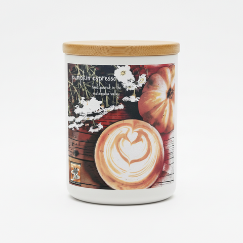 A matte white glass jar candle with a light bamboo lid, featuring a printed autumn-themed label that reads 'pumpkin espresso' and shows a pumpkin, white flowers, and a latte art heart design and the text 'hand poured in the matanuska valley