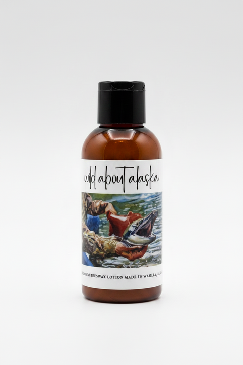 Trial size  'wild about alaska' premium beeswax lotion, featuring a central illustration of a person catching a salmon over water, with ingredients listed on the right and a 'Made in Alaska' seal on the left, all on a white background.