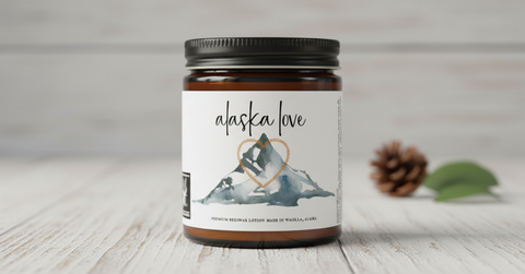A jar of 'alaska love' beeswax lotion stands on a light wood surface with a pinecone and green leaves blurred in the background. The product is in an amber glass jar with a black lid and a white label featuring a mountain graphic and a gold heart outline.