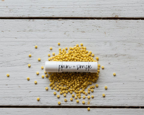 A white cylindrical cosmetic tube labeled 'plain + simple' stands upright on a black surface scattered with small yellow beeswax pellets, with a blurred wall in the background.