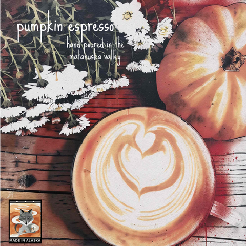 A square product label featuring a cup of latte art coffee on a rustic wooden table, with a whole pumpkin, white flowers, and overlaid text reading 'pumpkin espresso' and 'hand poured in the matanuska valley.' A small 'MADE IN ALASKA' label with a fox logo appears in the lower left.