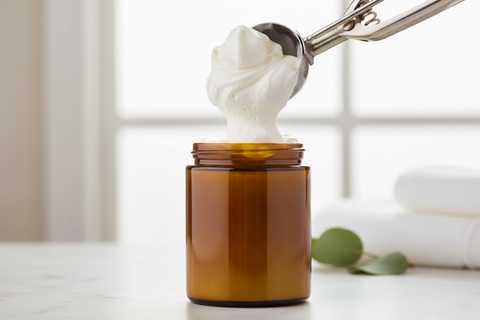 Rich white beeswax lotion being scooped from an open amber glass jar, with soft towels and greenery in the background.