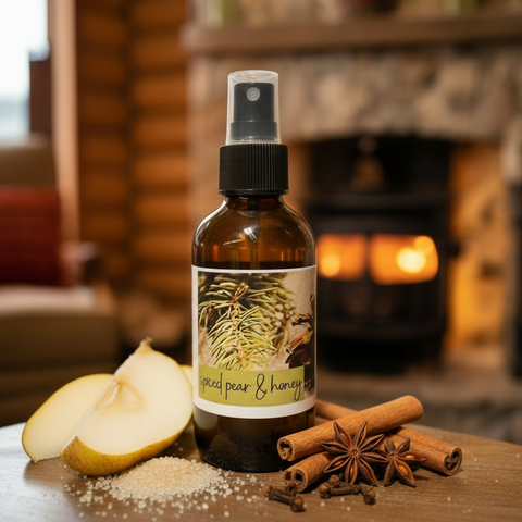 An amber glass spray bottle labeled 'spiced pear & honey' stands on a wooden surface in front of a warm fireplace. Sliced pears, cinnamon sticks, star anise, and cloves are arranged around the bottle as styled props.