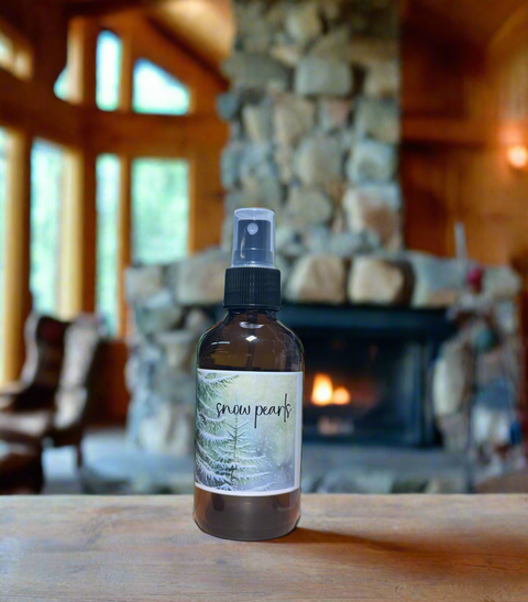 Snow Pearls linen and room mist in amber spray bottle with snowy spruce tree label, on a wooden table inside a cozy Alaska log cabin with stone fireplace glowing in the background, Grey Fox Candles"