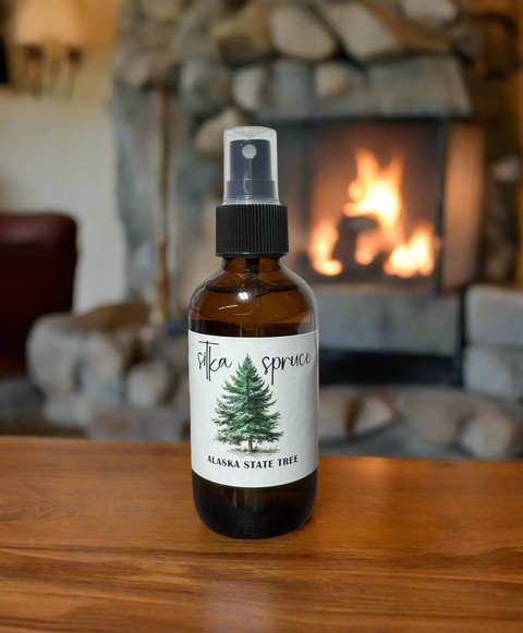 An amber glass spray bottle labeled 'Alaska State Tree' Sitka Spruce' sits on a wooden table in front of a blurred stone fireplace with a fire burning inside.