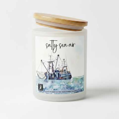 A frosted glass jar candle with a light wood lid, featuring a watercolor label of a fishing boat on the sea with the scent name 'salty sea air'.