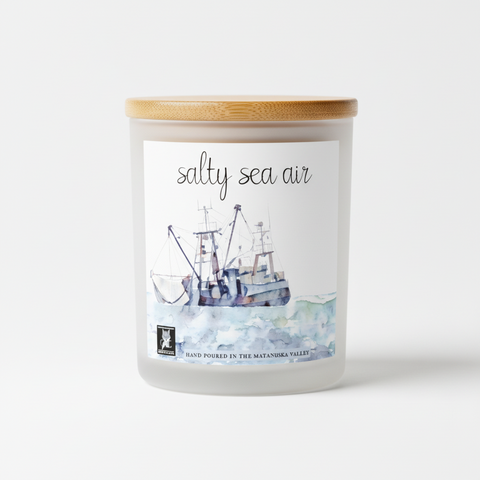 A square watercolor-style candle label with the words 'salty sea air' at the top and a painted fishing boat on a white background. Text at the bottom reads 'HAND POURED IN THE MATANUSKA VALLEY'.