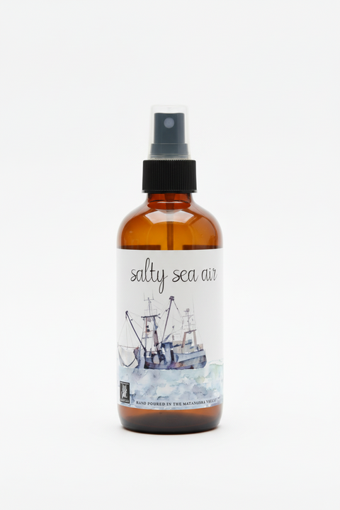 An amber glass spray bottle labeled 'salty sea air' with a black spray top and a watercolor-style label showing fishing boats on the ocean, photographed on a clean white background.