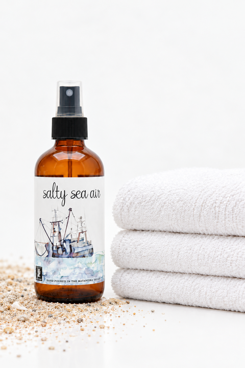 An amber glass spray bottle labeled 'salty sea air' with a black spray top, placed on a white surface with scattered coarse sand and three folded white towels beside it.