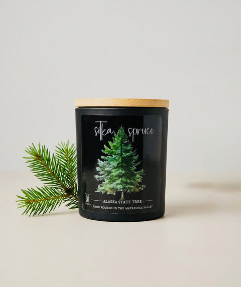 A matte black jar candle with a bamboo wood lid sits on a light beige surface next to a pine branch. The front label features a green pine tree illustration and the scent name 'SITKA SPRUCE'.