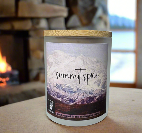 Candle labeled 'summit spice' on a wooden surface with a fireplace and window in the background