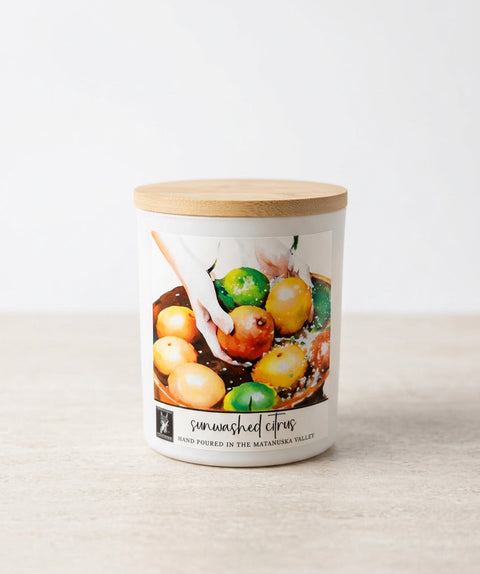 A matte white glass candle jar with a bamboo wood lid, featuring a label that reads 'sun-washed citrus' and shows a photo of citrus fruits. The candle is set on a light beige surface against a white background.