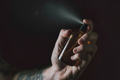 A hand holding and spraying a small amber glass bottle with a black spray nozzle against a dark background.