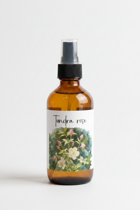 An amber glass spray bottle with a black spray top and a white label featuring the text 'Tundra rose' and a botanical illustration of white flowers and green leaves, photographed on a clean white background.
