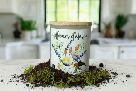 A frosted glass jar candle with a light bamboo lid sits on a white table in a softly blurred kitchen setting. The front label reads "wildflowers of alaska" and features colorful wildflower illustrations and a small bird graphic.
