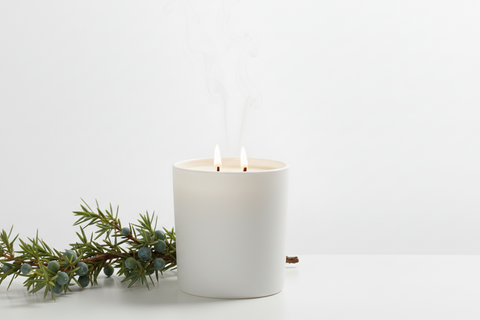 A white double-wick candle in a matte white glass jar with two lit wicks, styled on a white surface next to a green sprig of juniper berries against a clean white background.