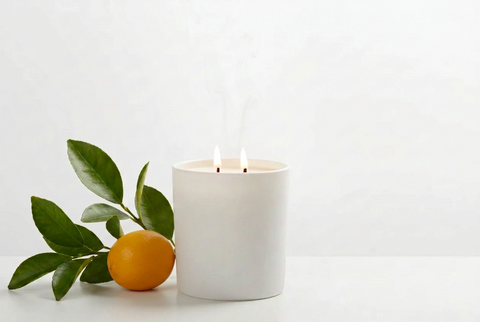 A white double-wick candle with two small flames and rising smoke, styled on a white surface next to green leaves and a small meyer lemon against a clean white background.