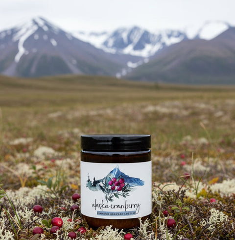 Jar of 'Alaska Cranberry' premium beeswax lotion with mountain and cranberry design on label, set against a natural landscape.