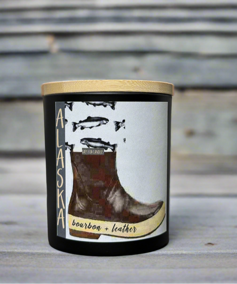 An 8oz matte black glass jar candle with a wooden lid, labeled with 'Alaska' and the scents 'bourbon + leather'. The label features an image of a boot and the candle is set against a rustic wooden backdrop.