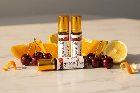 warm tones, cherries and citrus on a marble surface, two small perfume roller bottles with gold caps and beautiful labels are arranged in cherry grenadine fragrance, made in alaska by grey fox candles.