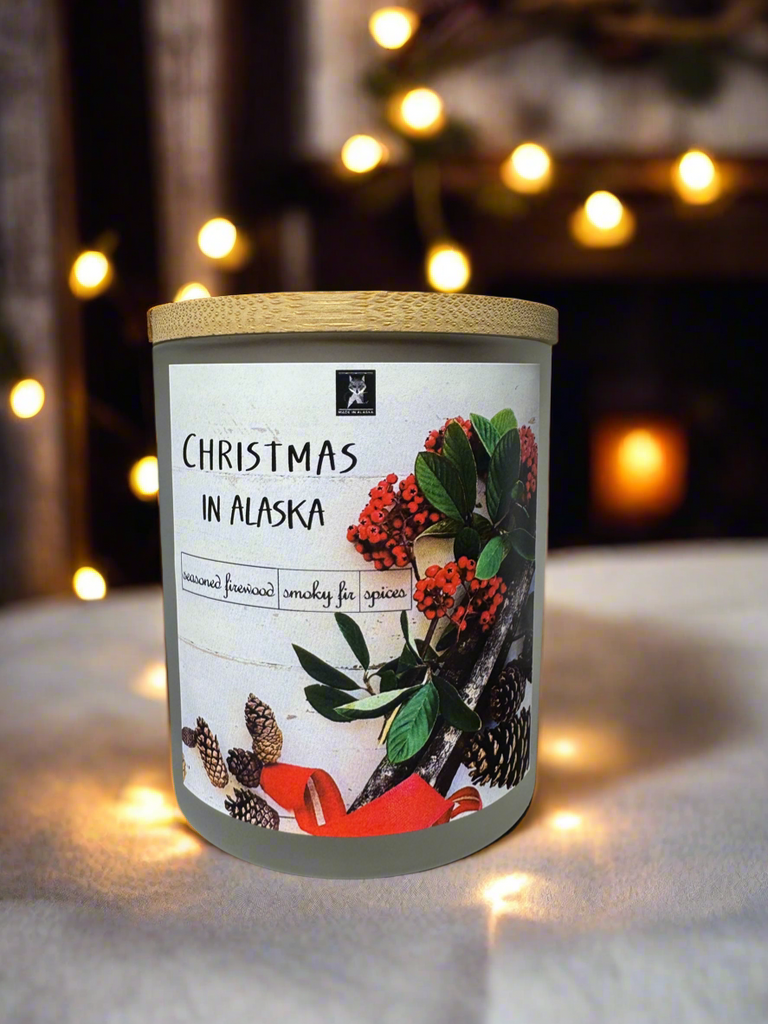 Candle with a label titled 'Christmas in Alaska' against a softly blurred background with lights.
