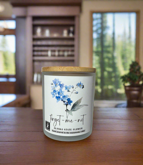 Candle with a floral label on a wooden table in a room with large windows.