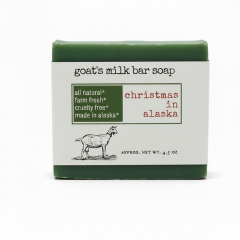 Product label for goat's milk bar soap with the scent 'christmas in alaska'. The label features a dark green box with white text listing key attributes, red text for the scent name, a black-and-white line drawing of a goat, and the net weight at the bottom.