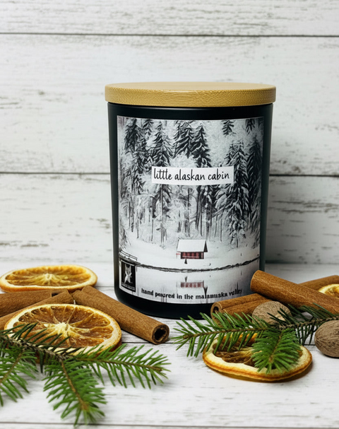 A black jar candle with a light wood lid and a winter forest label reading 'little alaska cabin,' styled on a white wood surface with cinnamon sticks, dried orange slices, pine branches, and small pinecones.