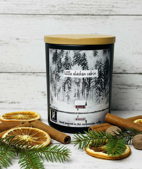 A jar candle with a bamboo lid and a winter forest label reading 'little alaska cabin,' styled on a white wood surface with cinnamon sticks, dried orange slices, pine branches, and walnuts.