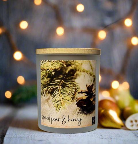 Candle with a label featuring pine branches and the scent 'spiced pear & honey' against a blurred background with lights and fruits.