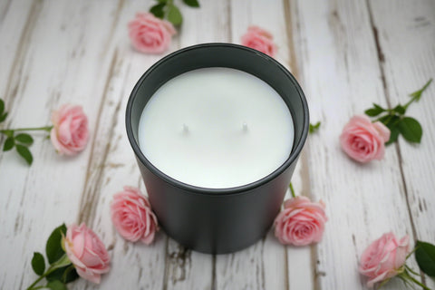 White candle in a black container on a wooden surface with pink rose flowers.