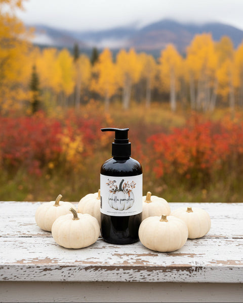 A black pump bottle with a white label reading 'vanilla pumpkin' and a pumpkin illustration, standing on a wooden farmhouse table with autumn trees in the background.